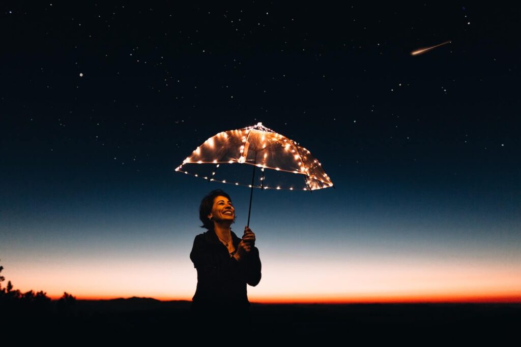 Person standing at night with an umbrella containing small lights and in the background is a red horizon, starts, and a comet.