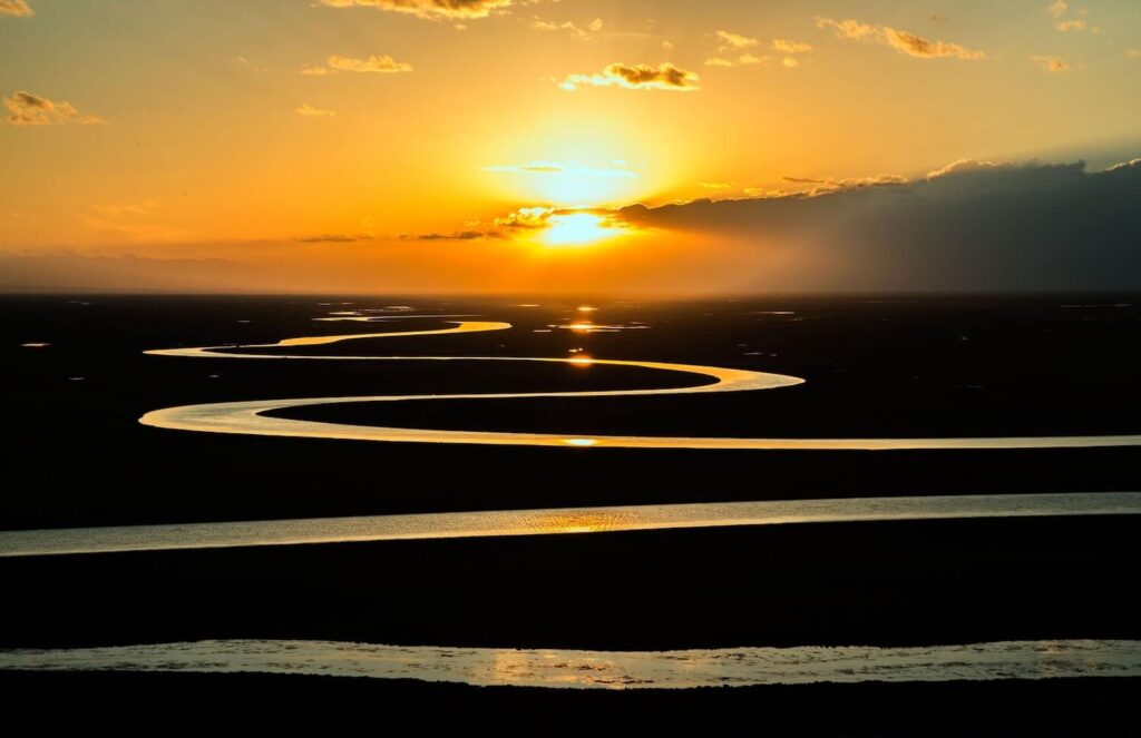 The sun setting through clouds with a twisting river running across a flat landscape