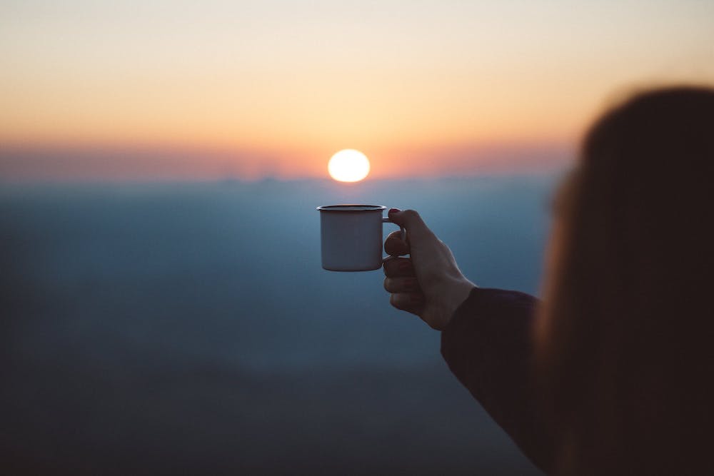 A persons' hand holding up a mug with a sunrise in the background